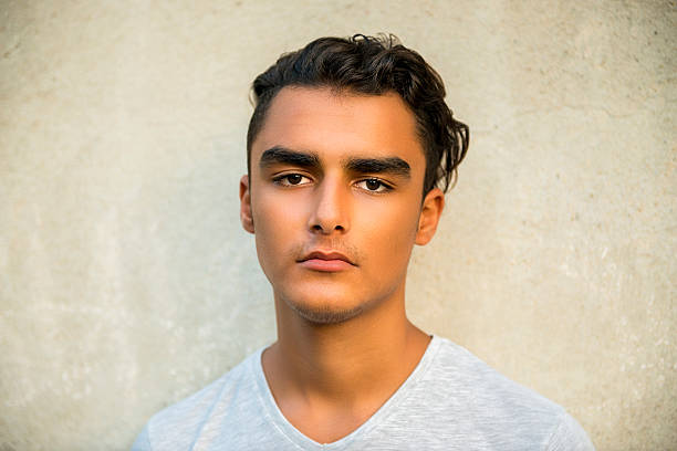 Portrait of young man looking at camera with blank expression. Horizontal composition. Studio shot. Young man standing in front of colored (white color) background and looking at camera over it. Image taken with NIKON D800 and developed from RAW format.