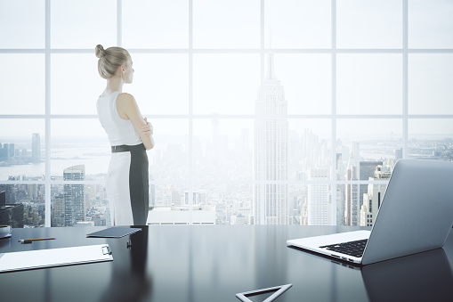 Thoughtful businesswoman standing in office with laptop, clipboard and other items on desktop. Window with New York city view in the background. 3D Rendering