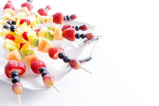 Serving of colorful healthy tropical fresh fruit kebabs with an assortment of exotic fruit on a white platter for a tasty vegan or vegetarian buffet, over white with copy space