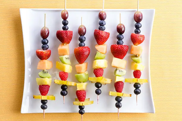 Healthy tropical summer fruit kebabs in a colorful arrangement on a modern white rectangular plate on a textured yellow table, overhead view
