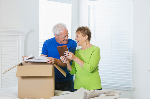 A senior couple moving house, packing or unpacking a cardboard box in the bedroom. The woman is holding a picture frame and they are looking at each other, smiling and talking.