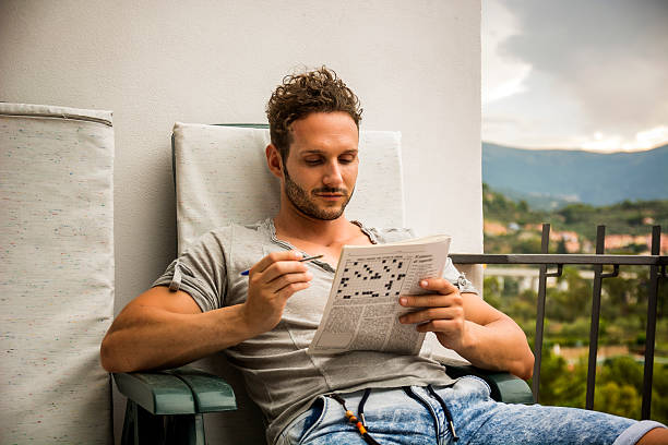 Young man sitting doing a crossword puzzle Young man sitting doing a crossword puzzle looking thoughtfully at a magazine, as he tries to think of the answer to the clue crossword stock pictures, royalty-free photos & images