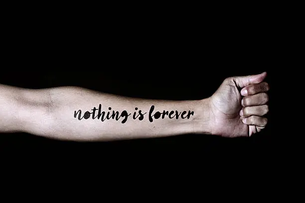 closeup of a young caucasian man with the text nothing is forever simulating a tattoo in his forearm, against a black background