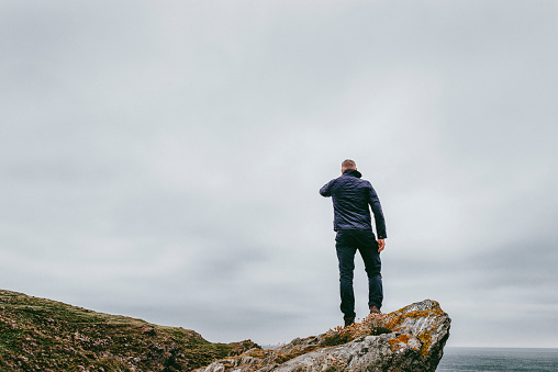 Man stood alone on a cliff top, looking out over a grey dramatic foreboding sky.