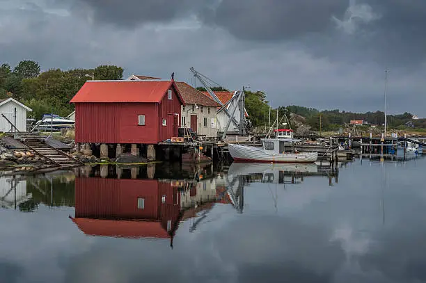 Old buildings and a dock on the Swedish island of South Koster are surrounded by small fishing boats.  The scene is reflected in the foreground.  The island is, with few exceptions, vehicle free.