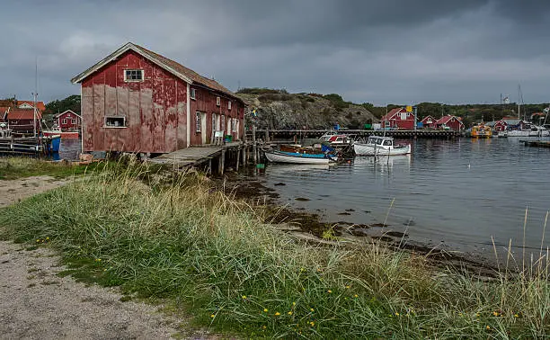 Old buildings and a dock on the Swedish island of South Koster are surrounded by small fishing boats.  The scene is reflected in the foreground.  The island is, with few exceptions, vehicle free.