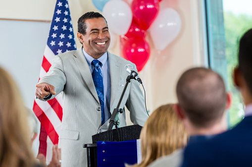 Mature adult African American man is smiling and pointing to an audience member while giving speech. Man is local politician who is standing behind a podium.