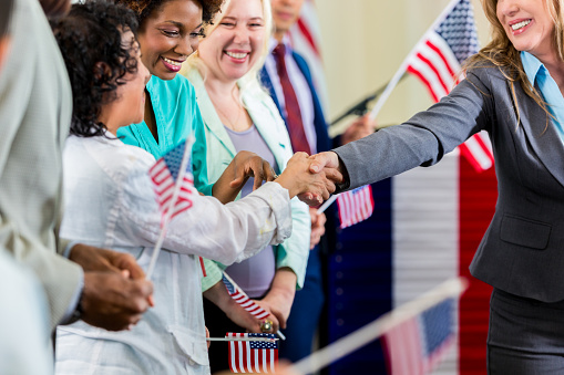 Senior adult Caucasian woman is local political candidate. She is smiling and shaking hands with diverse supporters during town hall meeting.