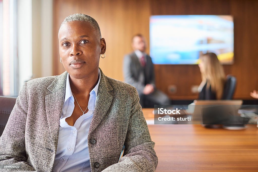 senior businesswoman Management team meeting in the boardroom. The team are sat round the conference table looking through at the companies performance figures for the month, a senior member of staff, possibly the business owner or director is sat smiling to camera as the meeting continues behind her. Businesswoman Stock Photo