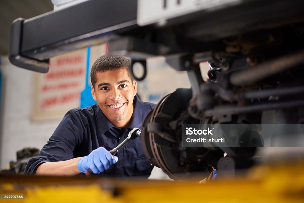 Brake discs A young mechanic is smiling to camera as he works on a car in a garage repair shop. He is wearing blue overalls.  He is fixing the brake discs on the front driver's side of the vehicle. Mechanic Stock Photo