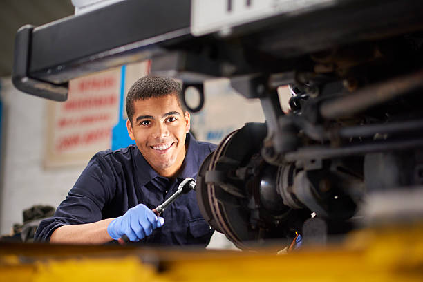 Brake discs A young mechanic is smiling to camera as he works on a car in a garage repair shop. He is wearing blue overalls.  He is fixing the brake discs on the front driver's side of the vehicle. auto mechanic photos stock pictures, royalty-free photos & images