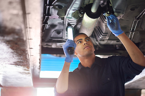 A young mechanic is working under a car in a garage repair shop. He is wearing blue overalls.  He is looking up with an inspection lamp to check the possible damage that has been sustained to the exhaust.