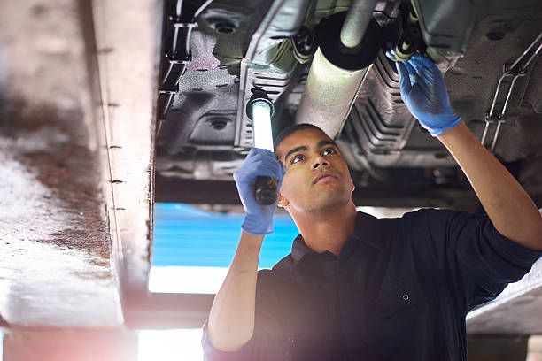 Mechanic checks exhaust A young mechanic is working under a car in a garage repair shop. He is wearing blue overalls. He is looking up with an inspection lamp to check the possible damage that has been sustained to the exhaust. mechanic working under car stock pictures, royalty-free photos & images