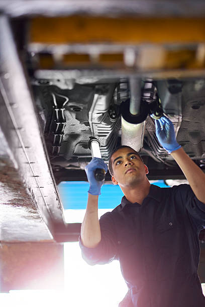 Exhaust repairs A young mechanic is working under a car in a garage repair shop. He is wearing blue overalls. He is looking up with an inspection lamp to check the possible damage that has been sustained to the exhaust. mechanic working under car stock pictures, royalty-free photos & images