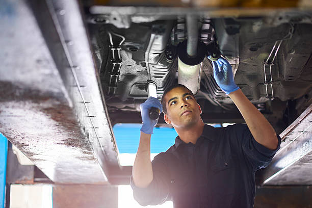 Auto mechanic A young mechanic is working under a car in a garage repair shop. He is wearing blue overalls. He is looking up with an inspection lamp to check the possible damage that has been sustained to the exhaust. mechanic working under car stock pictures, royalty-free photos & images