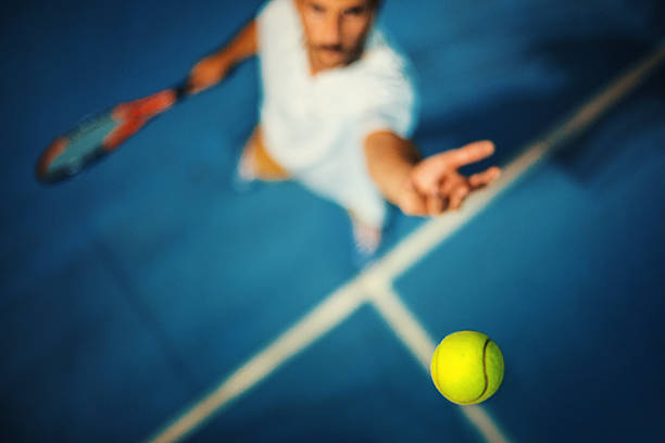 Tennis serve. Closeup top view of a male tennis player hitting a ball during a serve. It's played on blue hard court surface just like at US open. Very shallow focus, only tip of the racket and the ball are in focus. serving sport stock pictures, royalty-free photos & images
