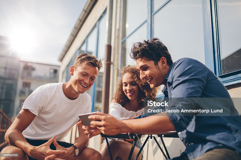 Group of friends watching video on smartphone Three young friends sitting outdoors and looking at mobile phone. Group of people sitting at outdoor cafe and watching video on the smartphone. Friendship Stock Photo