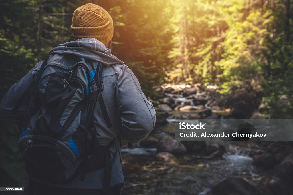 Traveler stands alone near the river in forest. Traveler walking alone in the mountain forest. Close-up portrait of a man with backpack, mountain river in front of him. Voyager - Spacecraft Stock Photo