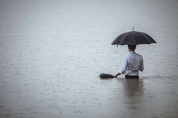 Handsome man standing in water and holding umbrella during rain Rear view on a handsome man wearing white shirt and holding umbrella and a briefcase, standing in the water on a rainy day. The man waded knee-deep in water. Selective focus, three quarter length, copy space has been left. walking in water stock pictures, royalty-free photos & images