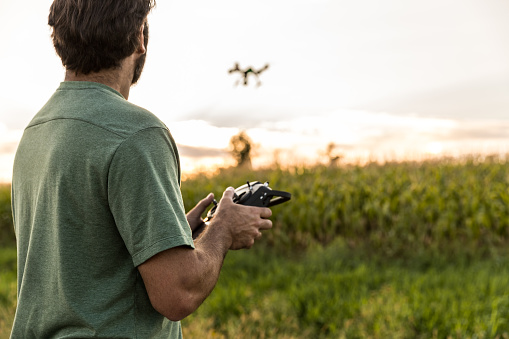 DSLR picture of a man flying a drone over a field by a beautiful summer sunset. The man is looking at the drone.