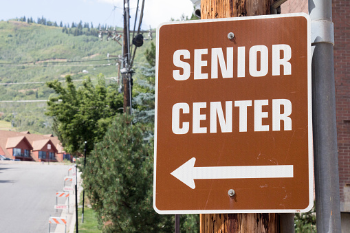 Image of a brown and white sign that says Senior Center with an arrow to the left.  The sign is pointing towards a distant blurry road and building.