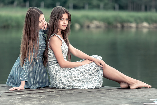 Two beautiful dark-haired girls teenagers posing together at the forest lake. The girls are sitting huddled together on a wooden quay. One of the girls looking at the camera. Other girl looking at the girlfriend. Shooting in the warm summer evening outdoors