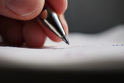 Male hand holding a sharp blue office pen and writing to the white lined notepad as a symbol of taking notes or business communication