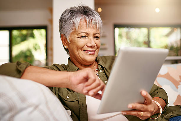 Staying in the the social loop thanks to technology Shot of a happy older woman relaxing on the sofa with her digital tablet at home old woman smiling tablet stock pictures, royalty-free photos & images