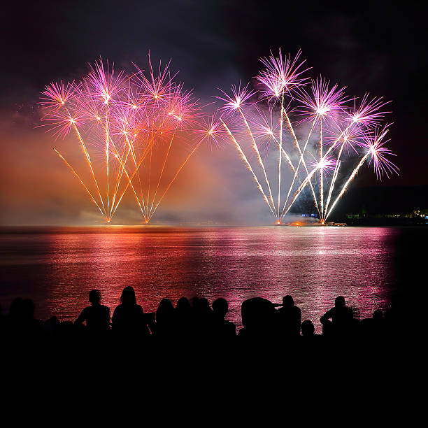 Square composition color photography at night of beautiful large and colorful fireworks display with awe pyrotechnic effects at edge of the water with some abstract reflections in water wave and with unrecognizable crowd of people silhouette watching, during celebration event like new year or national day. The original picture was taken during the Bastille Day in France, 14th of July, along Rhone river in Isere department in Auvergne-Rhone-Alpes region in Europe in Montalieu-Vercieu city.