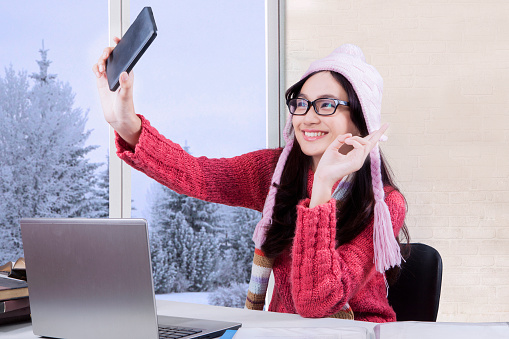 Female high school student taking selfie photo with a mobile phone while studying with laptop at home