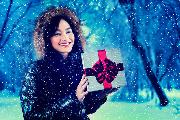 Cheerful teenage girl holding a gift box while smiling at camera and wearing winter coat, shot outdoors