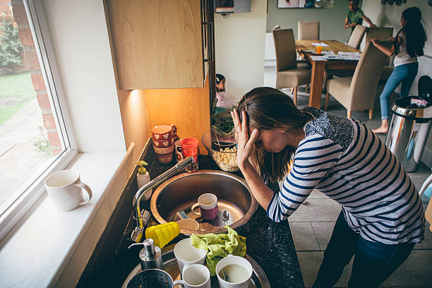 Stressed mother of three Stressed mum at home. She has her head in her hands at a messy kitchen sink and her children are running round in the background. unhygienic stock pictures, royalty-free photos & images