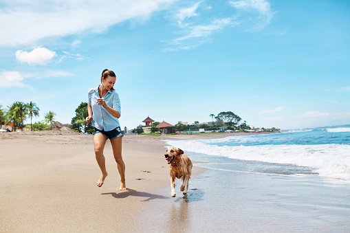 Summer Fun On Beach. Beautiful Happy Woman Running With Her Dog, Golden Retriever, Pet On Wet Sand In Sea Water. Holidays Vacations At Tropical Resort. Summertime Travel Concept.