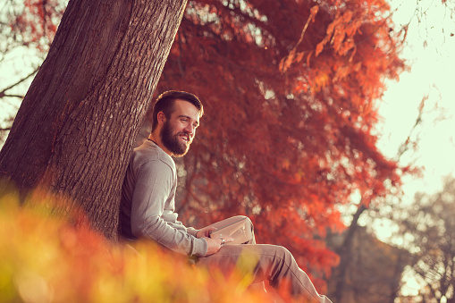 Handsome young man sitting on a fallen autumn leaves in a park, reading an e-book on a tablet computer