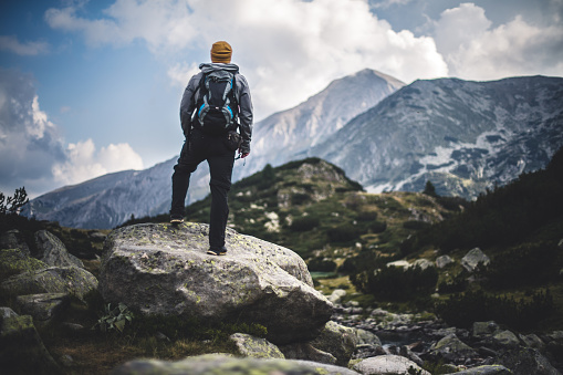 Traveler with backpack stands on a huge rock and looks on a mountain peak in front of him.