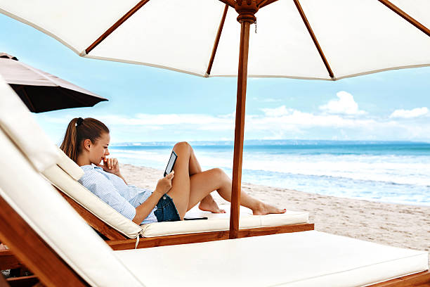 A woman is sitting on a beach chair reading a book under an umbrella. The scene is peaceful and relaxing, with the sound of waves in the background. The woman is enjoying her time alone