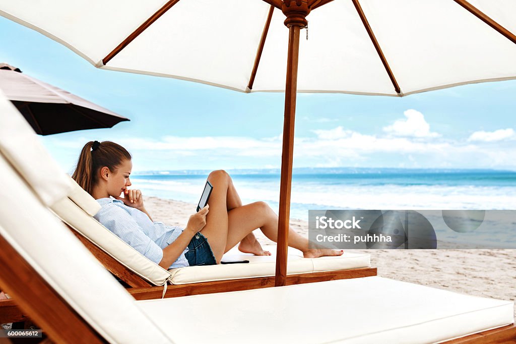 Summer Relaxation. Woman Reading, Relaxing On Beach. Summertime Summer Relaxation. Beautiful Woman Reading E-Book, Relaxing On Sun Lounger, Deck Chair Under Umbrella, Tent On Beach By Sea. Summertime. Holiday Vacations. Leisure, Recreation, Enjoyment Concept Beach Stock Photo