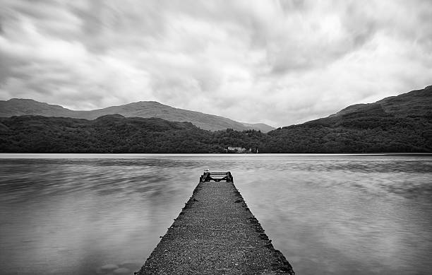 Long exposure and black and white image showing a stone pier jutting out into the waters of Loch Lomond, on the edge of the Scottish Highlands.