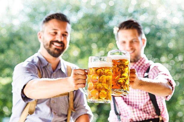 Men in traditional bavarian clothes holding mugs of beer Two handsome hipster young men in traditional bavarian clothes holding mugs of beer, clinking. Beer Fest. Sunny summer garden. Lederhosen stock pictures, royalty-free photos & images