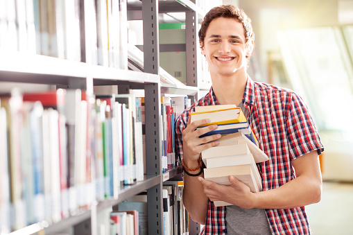 Portrait of a young student holding books standing by bookshelf in library