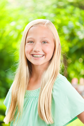Portrait of a happy smiling caucasian young teen blonde girl. She is wearing orthodontic dental teeth braces, looking at the camera. Photographed in head and shoulder portrait in outdoor setting with soft focus greenery in the background. Framed in vertical format.