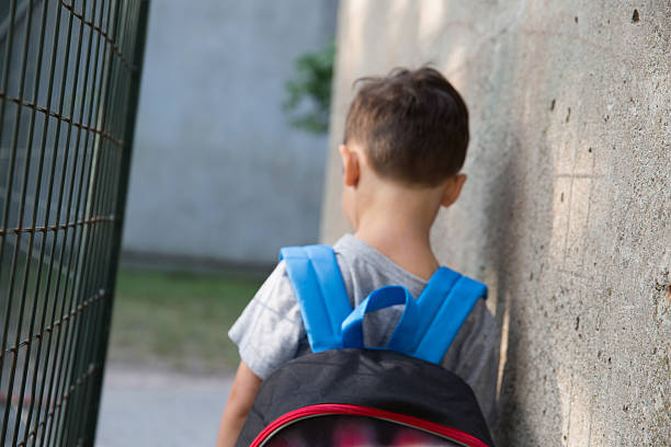 Alone in the Schoolyard Little boy slightly out of focus and blurred with his back to the camera wearing a backpack in the schoolyard school exclusion stock pictures, royalty-free photos & images