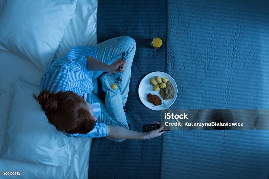 Woman and her life in a bed Photo from the top of young girl sitting on a bed, eating dinner and watching television Night Stock Photo