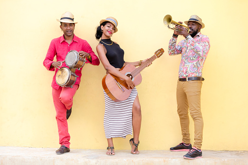 Cuban musical band, the trio consisting of a well known musicians standing against the bright yellow wall. Beautiful young woman standing in the middle, holding a guitar. The man on the left holding the small drums bongos, and a musician on the left holding a trumpet. Havana, Cuba, 50 megapixel image.