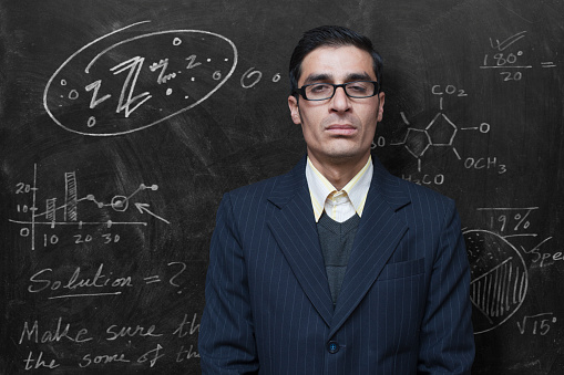 Indoor image of an exhausted male teacher or professor feeling sleepy in the classroom. He is standing against blackboard where his thought bubble is drawn depicting his sleepiness condition. The bespectacled man is in formal shirt, sweater and coat. One person, waist up, horizontal composition with copy space.