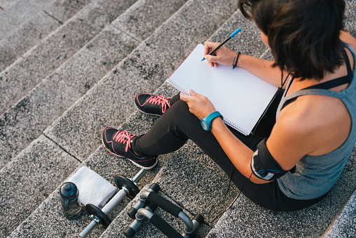 Fitness sporty woman writing on blank notepad while sitting on urban stone stairs before exercises workout routine. Female athlete focusing on her goals.