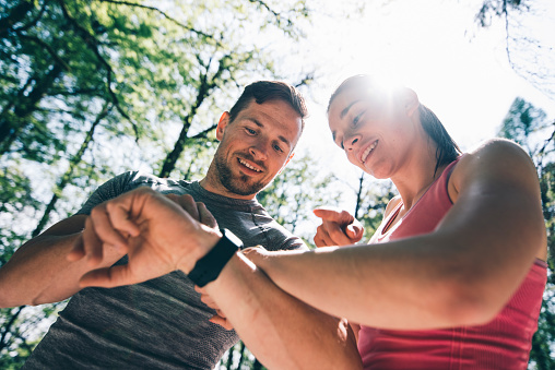 Young couple after training in nature. They are checking their smart watches after workout and discussing the results. Green trees are in back.