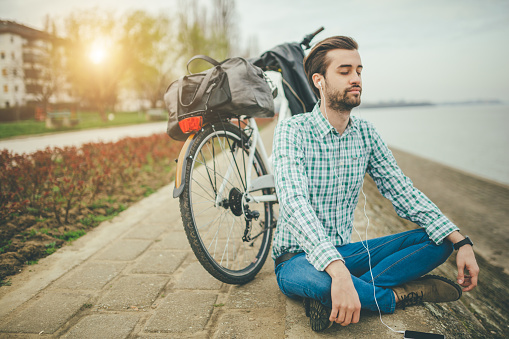 Young man is relaxing by the river. He is looking at the phone and smiling. He wears green shirt and blue jeans. He is sitting next to his parked bike.