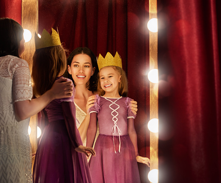 Cute little actress. Young mother and her daughter child girl in Princess costume on the background of theatrical scenes and mirrors.