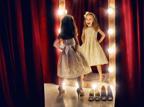 Cute little fashionista. Happy child girl try on outfits and mom's shoes looking at mirror.